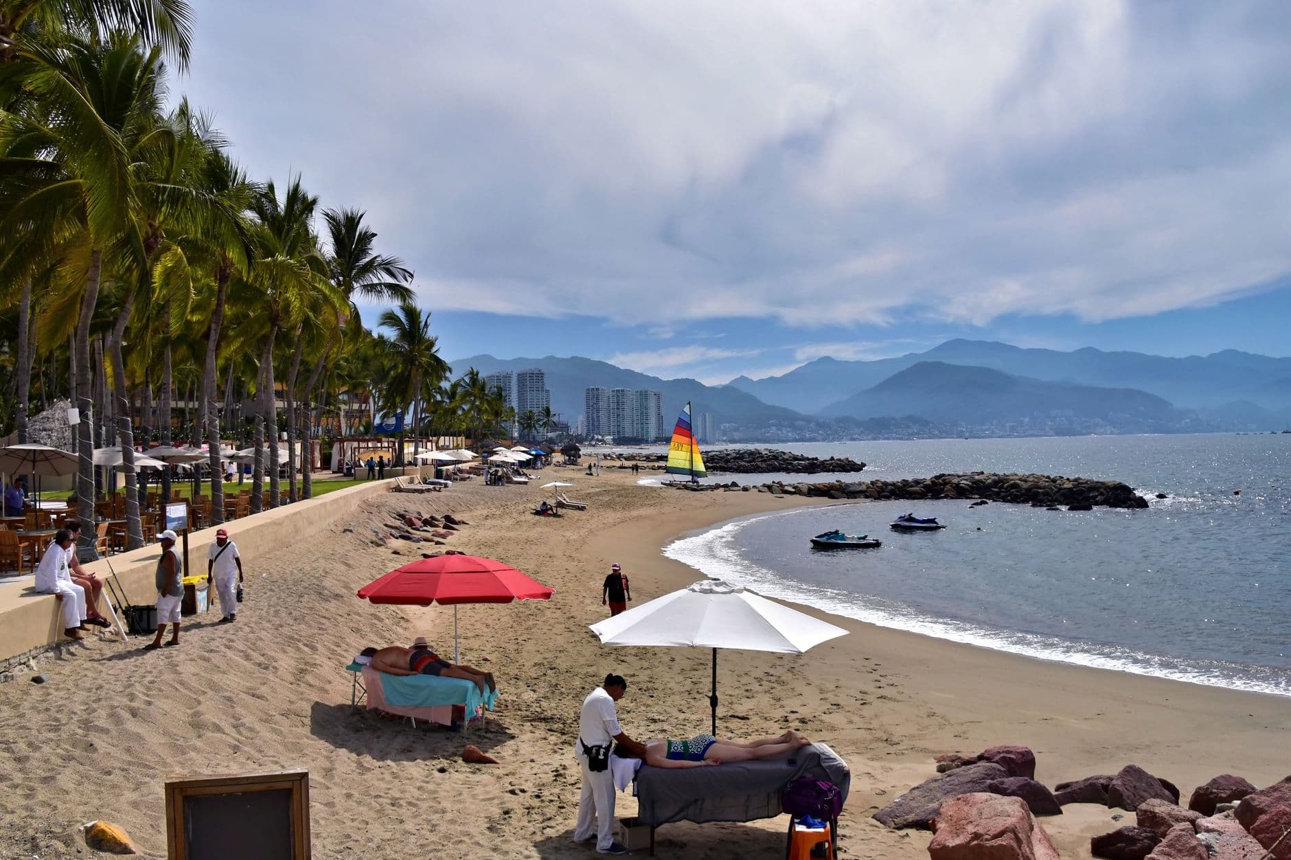 A calm Puerto Vallarta beach with palm trees, shade umbrellas, and a sailboat offshore
